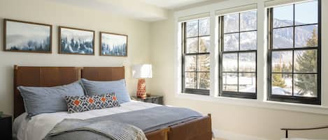 A bedroom with a large bed, a nightstand, a lamp, and a chair. There are three framed pictures on the wall, and a window with a view of mountains.