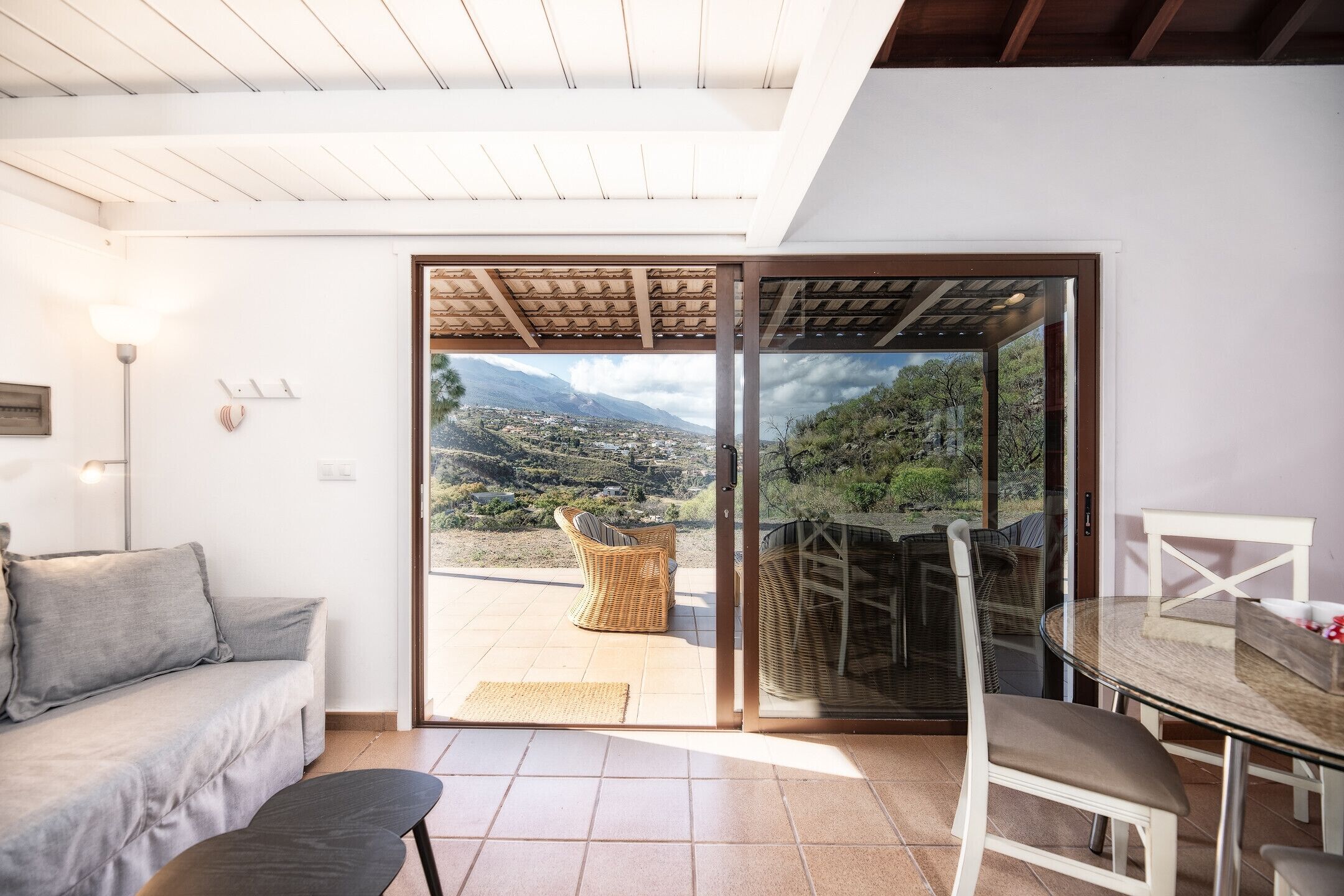 Uma sala de estar moderna com um sofá, uma mesa de jantar com cadeiras e uma porta de vidro deslizante que dá para uma varanda com vista para uma paisagem montanhosa.