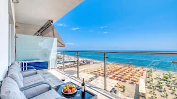 Un balcone con una divano, un tavolo con una ciotola di frutta e bicchieri da vino, e vista su una spiaggia con ombrelloni e lettini.