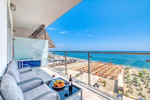 Un balcón con sofá, una mesa con fruteros y copas de vino, y vista a una playa con sombrillas y sillas de descanso.