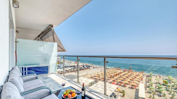 Un balcon avec un canapé, une table garnie de fruits et de vin, et une vue sur une plage parsemée de parasols et de chaises longues.