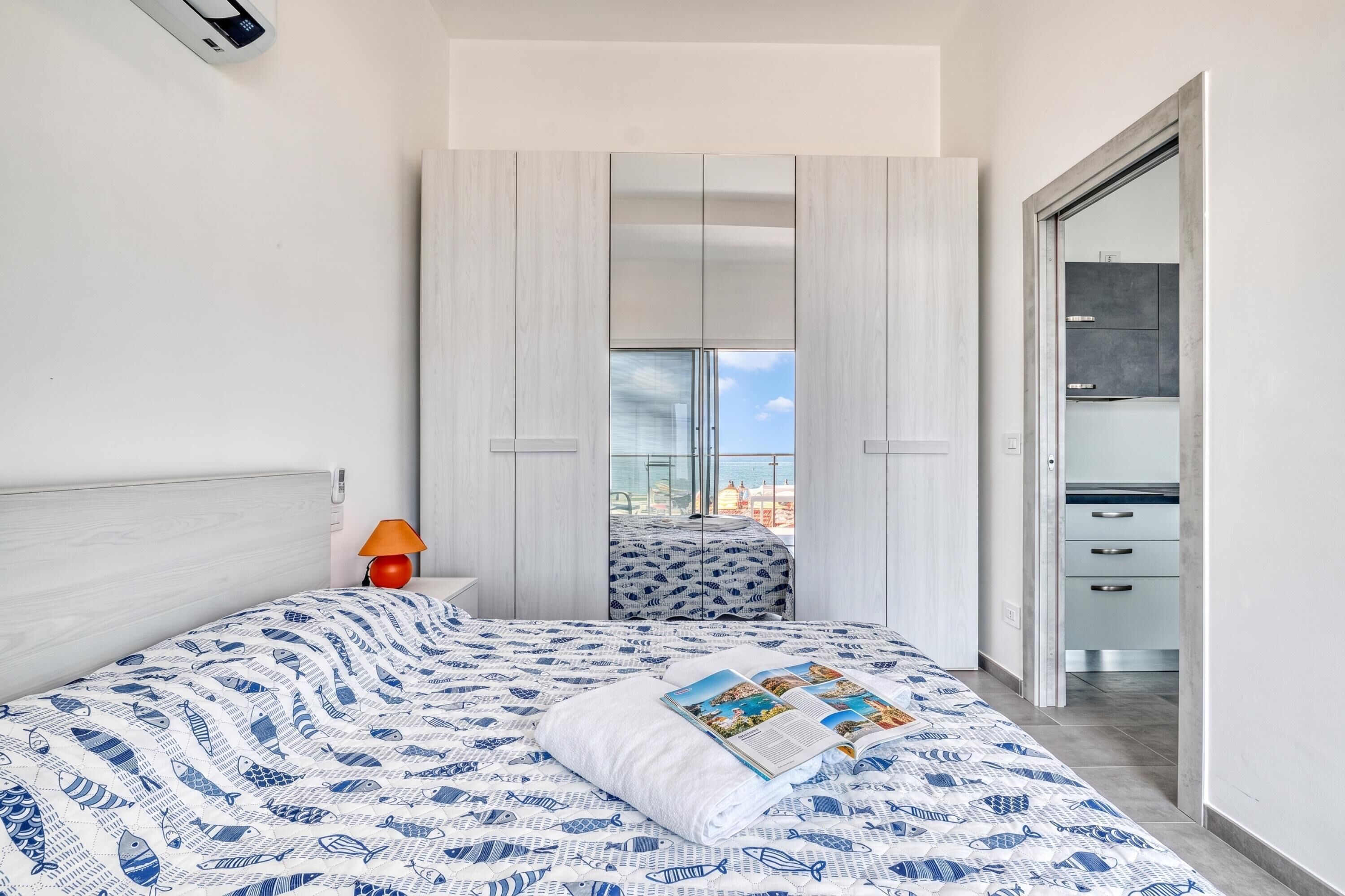 A modern bedroom with a bed, a nightstand, a lamp, and a view of the city through a large window.