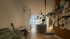A living room with a sofa, a piano, and a guitar. There is a kitchen area with stools and a counter. The room has a tiled floor and a wall with papers and photos.