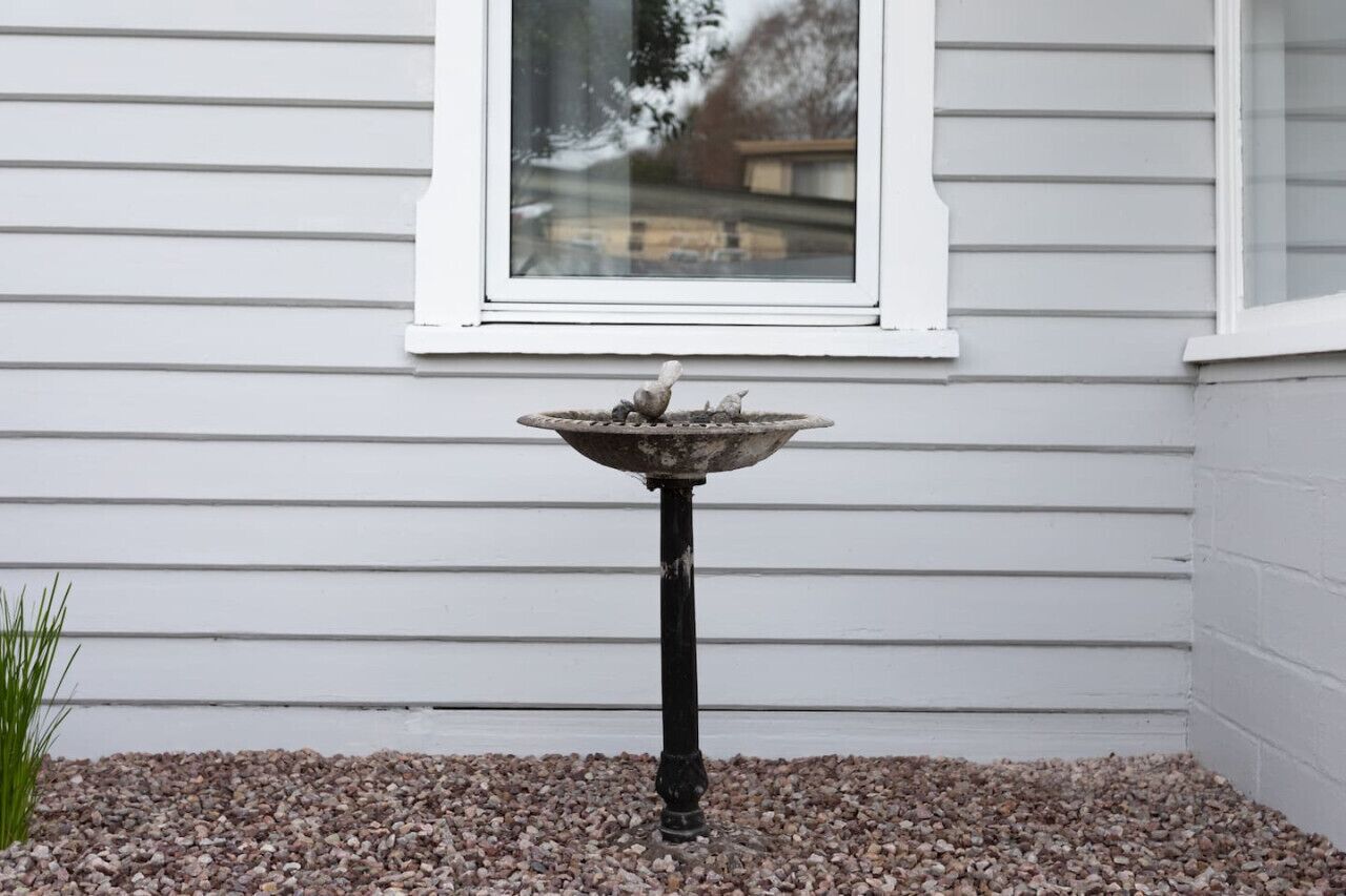 A bird bath on a pedestal with a gravel base, positioned in front of a white window with horizontal siding.