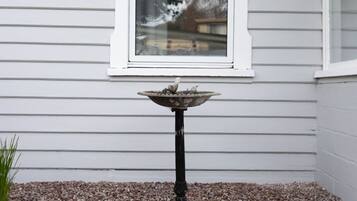 A bird bath on a pedestal with a gravel base, positioned in front of a white window with horizontal siding.