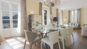 Un comedor con una mesa de vidrio, sillas blancas y una araña. Se observa un sofá y un televisor al fondo.