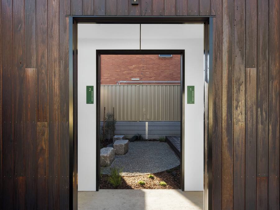 Entrada de un edificio moderno con fachada de madera, un camino de hormigón y una zona de grava con piedras.