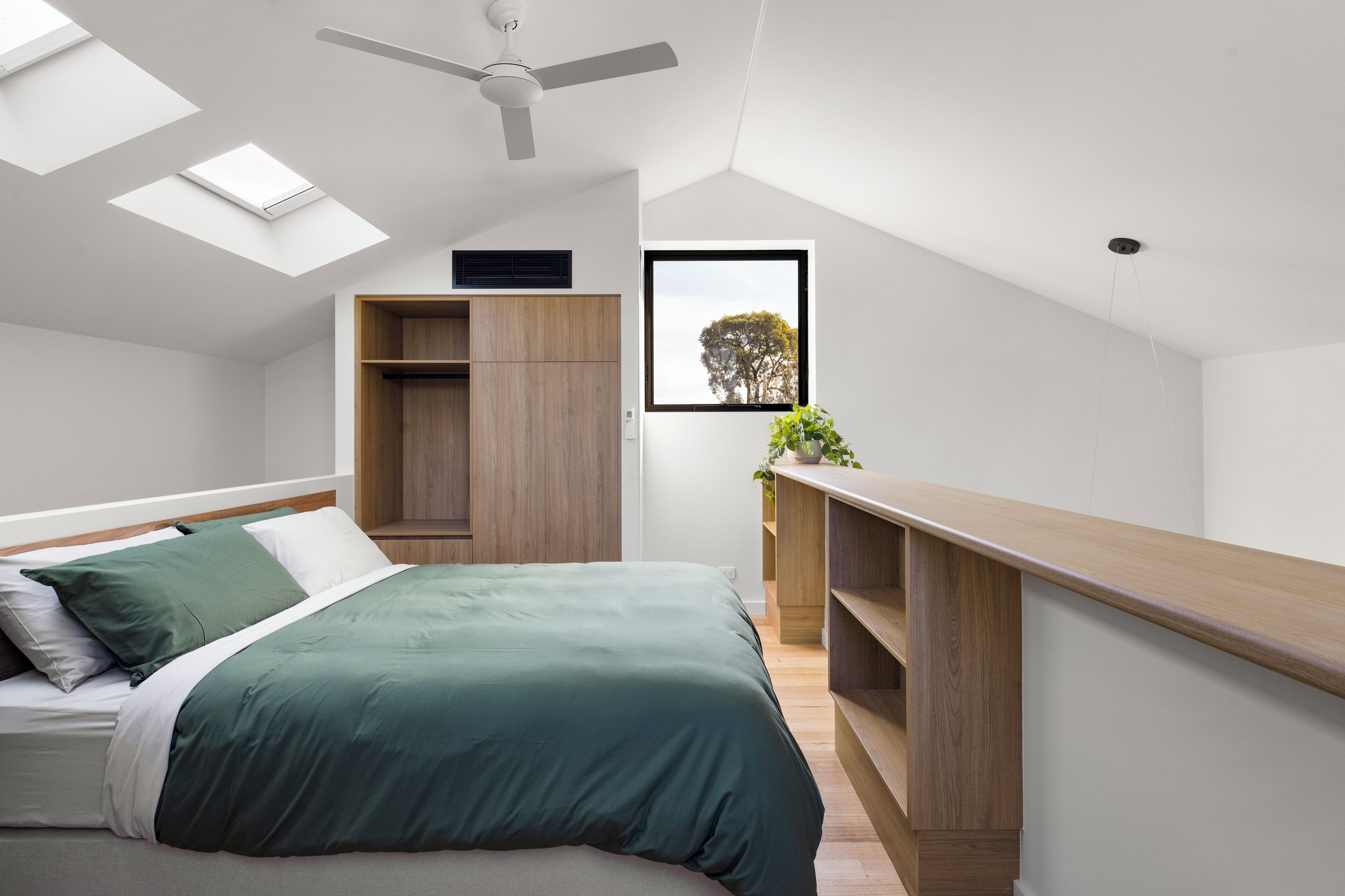 Un dormitorio moderno con tragaluz, una cama con ropa de cama verde, un armario de madera y vistas a árboles a través de una ventana.