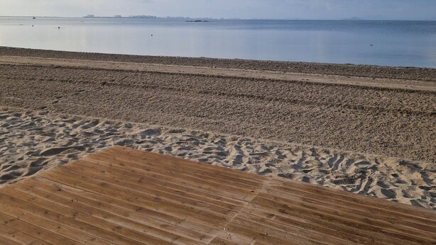 A beach with a wooden boardwalk leading to the water, a calm sea, and a clear sky with scattered clouds.