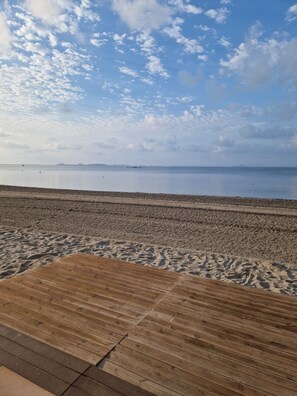 A beach with a wooden boardwalk leading to the water, a calm sea, and a clear sky with scattered clouds.