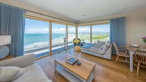Ein großzügiger Wohnbereich mit einem großen Fenster zum Meer, eine gemütliche Sitzecke mit Sofa und Sesseln sowie ein Esstisch mit Stühlen.