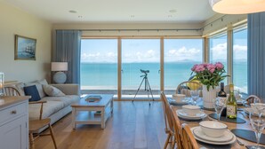 Ein geräumiges Wohnzimmer mit großem Fenster und Meerblick, ein Essbereich mit Tisch für zwei Personen sowie eine gemütliche Sitzecke mit Sofa und Lampe.