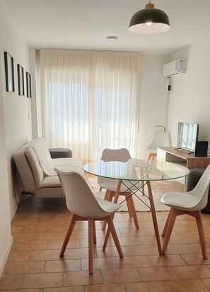 Un salon moderne avec une table à manger en verre, des chaises blanches, un canapé et un luminaire suspendu au plafond.