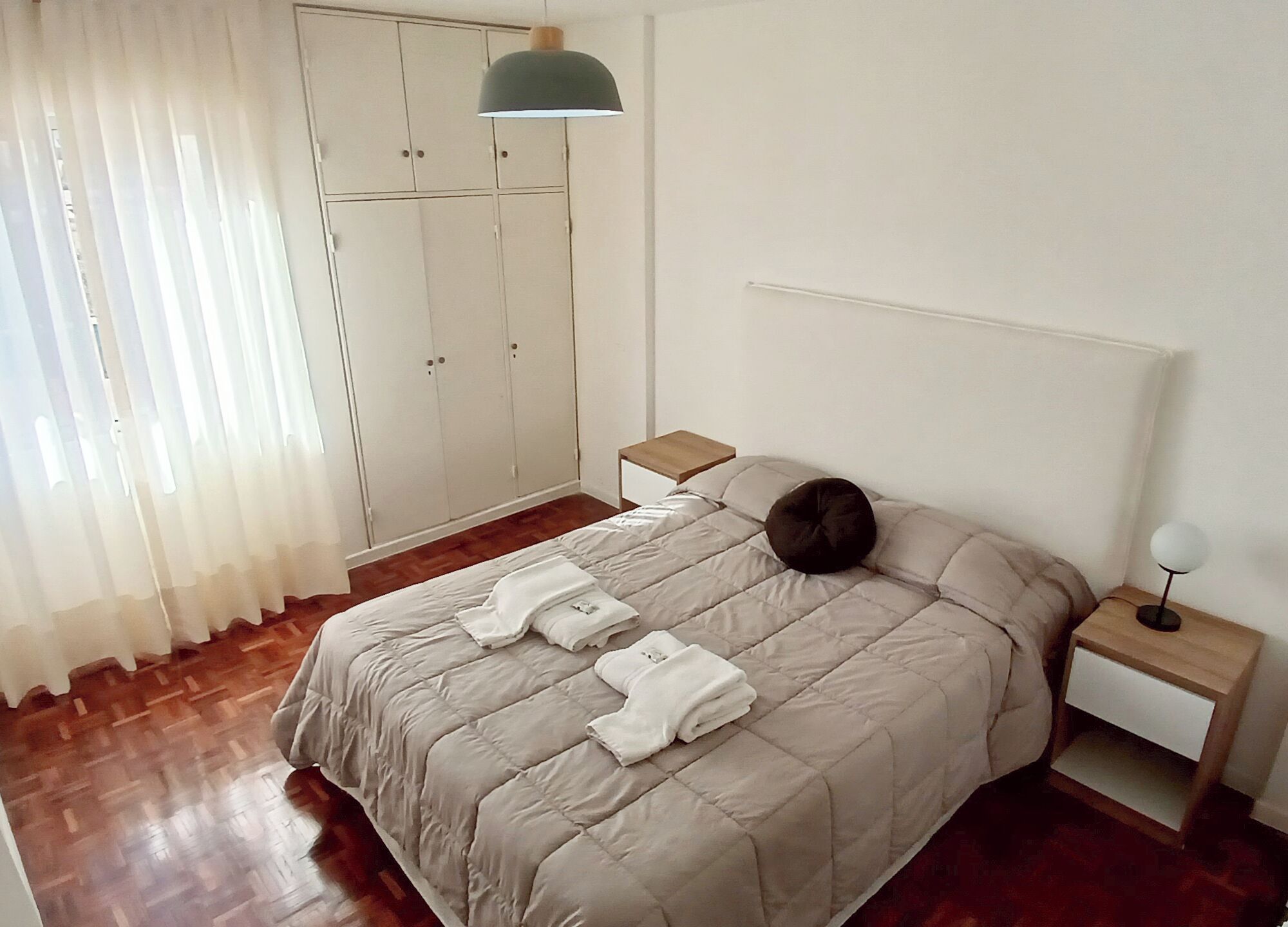 Een net opgemaakt bed met een beige dekbedovertrek, twee witte handdoeken, een nachtkastje met een lamp en een garderobe met kledingroede.