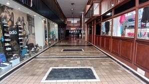 A corridor with a tiled floor, glass storefronts on one side, and a wooden railing on the other. - Tempo Corrientes (Córdoba)