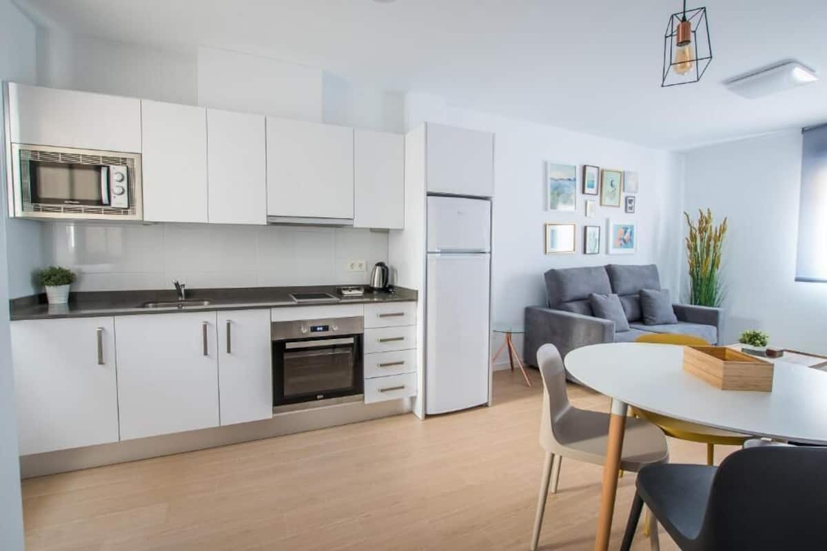 A modern kitchen with white cabinets, a microwave, and a refrigerator. There is a dining area with a white table and chairs.