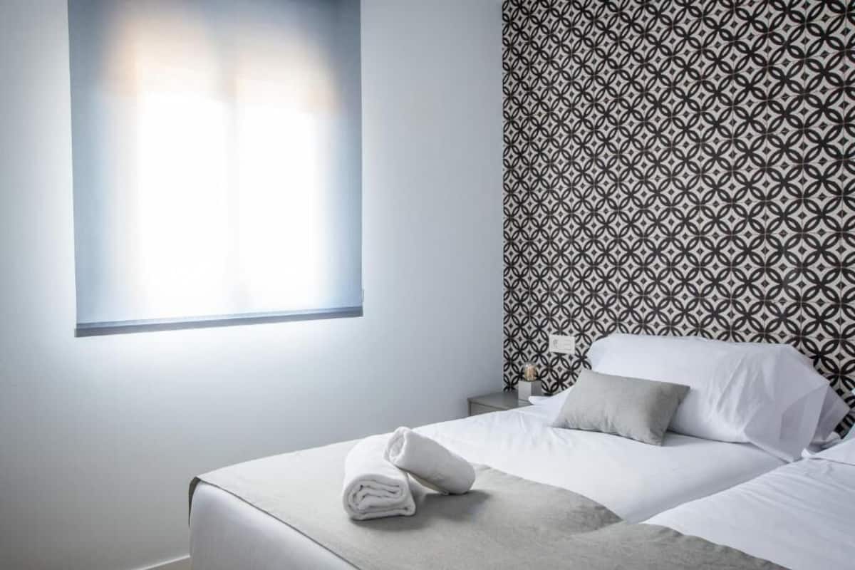 A hotel room with a bed, a nightstand, a window with blinds, and a patterned wallpaper.