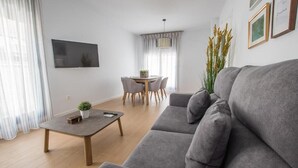 A modern living room with a grey sofa, a coffee table, and a dining table with chairs. There is a TV mounted on the wall and a plant on the coffee table. - Ibericabeds Granada (Granada)