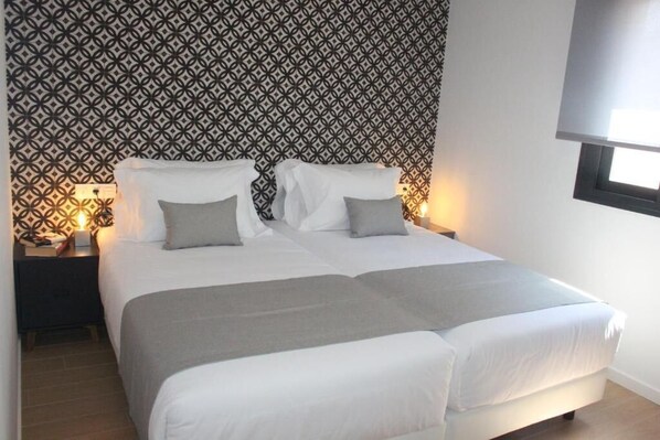 A double bed with white and gray bedding, a patterned wallpaper, and a nightstand with a lamp. - Ibericabeds Granada (Granada)