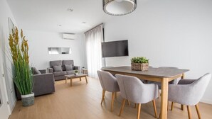 A modern living room with a grey sofa, a dining table with four chairs, a TV mounted on the wall, and a large potted plant. - Ibericabeds Granada (Granada)