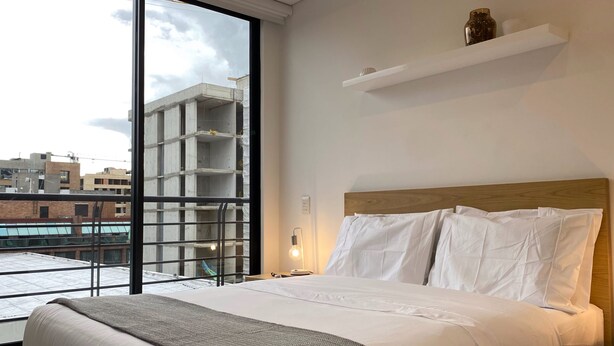 Ein Hotelzimmer mit großem Fenster, einem Bett mit weißer Bettwäsche, einem Holz-Kopfschild und einem weißen Regal mit Dekorationen.