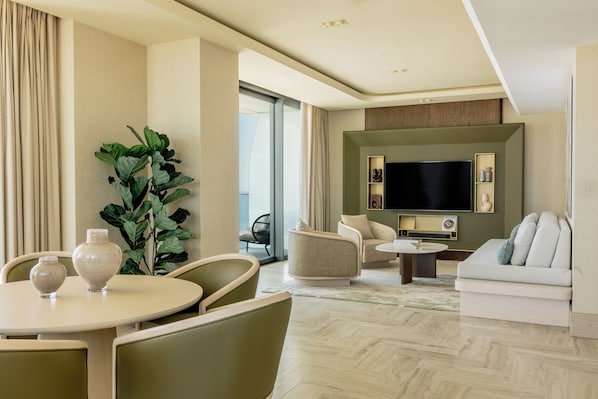 Una habitación de hotel moderna con un televisor de pantalla plana grande, un sofá, una mesa de comedor con sillas y una planta grande.