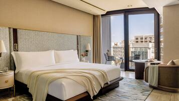 Habitación de hotel con una cama grande, una mesita de noche, una silla, una pequeña mesa y vistas a la ciudad a través de un ventanal amplio.