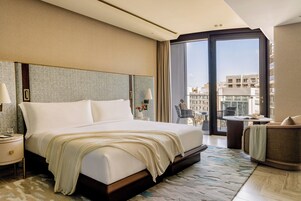 Habitación de hotel con una cama grande, una mesita de noche, una silla, una pequeña mesa y vistas a la ciudad a través de un ventanal amplio.