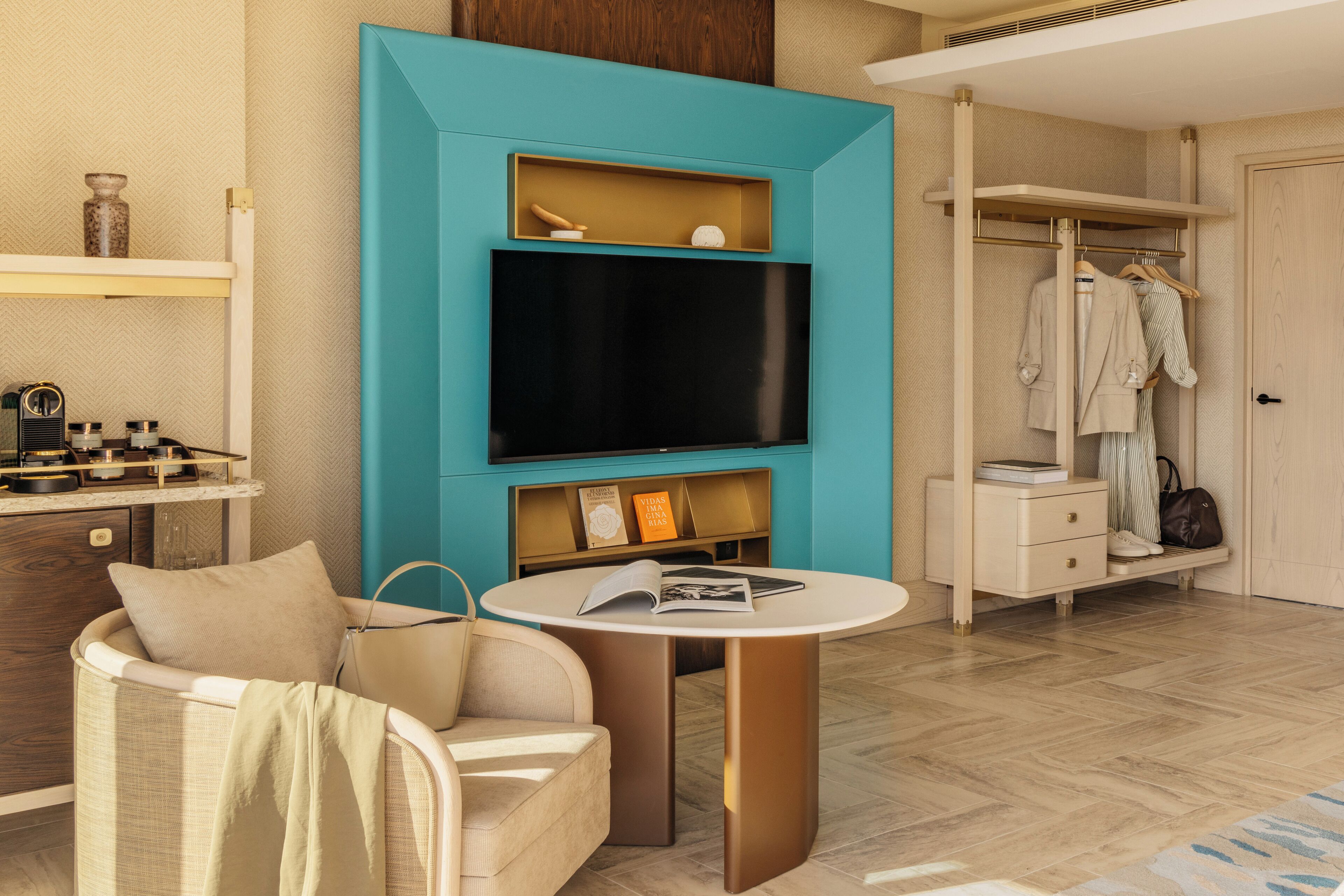 Une chambre d’hôtel moderne avec un mur d’accent de couleur turquoise, un téléviseur à écran plat, une table ronde avec des livres, un canapé, une armoire et une coiffeuse avec un miroir.