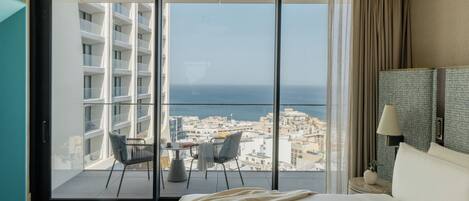 A hotel room with a bed, a nightstand, a lamp, a chair, a small table, and a view of the city and sea through large glass doors.