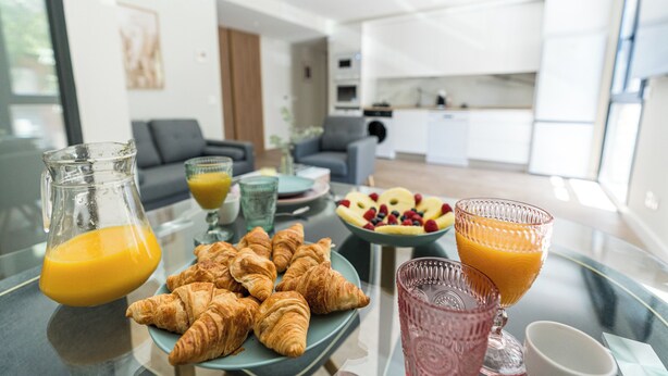 Une table de petit-déjeuner avec des croissants, des fruits et du jus d’orange, dans un espace de vie moderne avec une cuisine en arrière-plan.