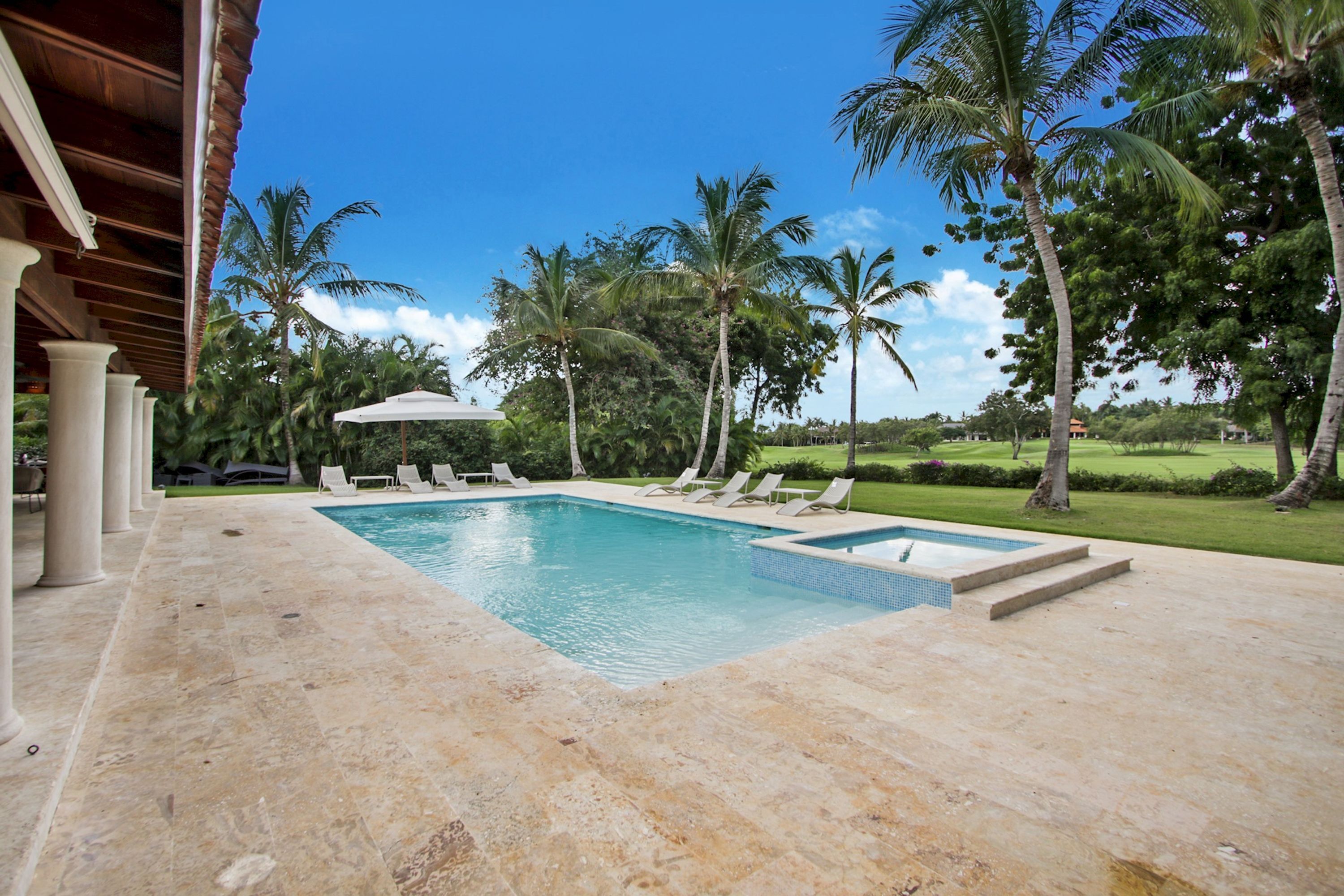 Piscina retangular com um deck ladrilhado, rodeada por palmeiras e espreguiçadeiras.