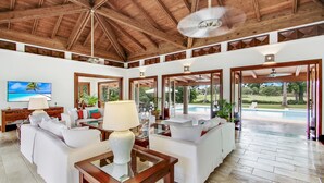 A spacious living room with white furniture, a wooden ceiling, and large windows offering a view of the outdoors. - Gorgeous Villa at Casa de Campo With Large Pool Chef Maid and 2 Golf Carts (La Romana)