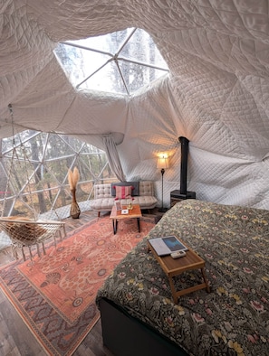 A cozy interior with a large bed, a sofa, a hanging chair, and a small table with a lamp.