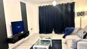 Una sala de estar moderna con un televisor de pantalla plana, una mesa de centro de vidrio, un sofá y una cortina oscura.
