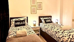 Una habitación de hotel con dos camas, cada una con una colcha estampada y almohadas con texto. Hay cuadros enmarcados en la pared, una lámpara de noche y una bombilla colgando del techo.