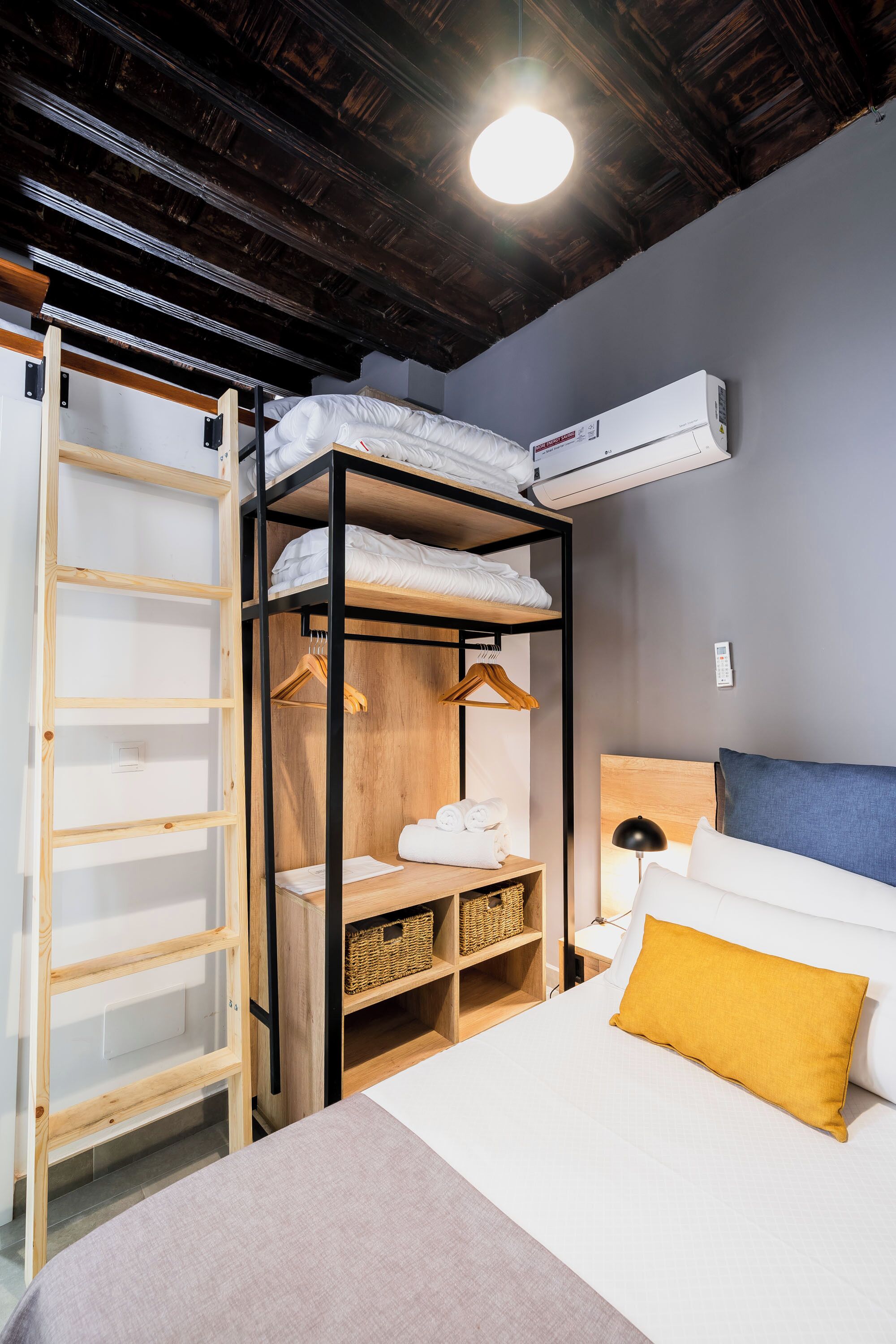 Un lit superposé avec une échelle en bois, une armoire, une table de chevet et un lit simple avec un oreiller de couleur moutarde.