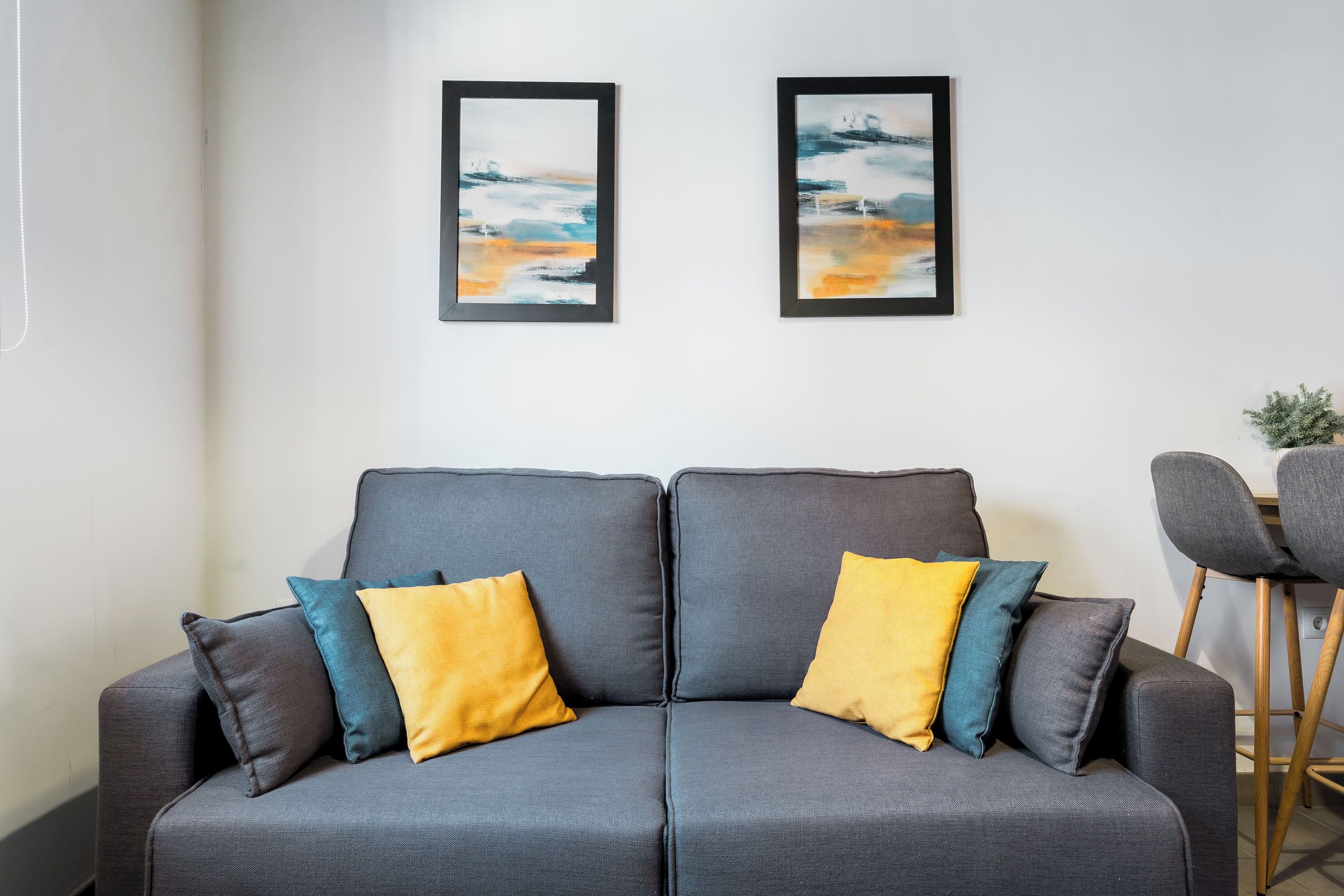A grey sofa with yellow and blue cushions, two framed abstract paintings on the wall, and a wooden chair in the background.