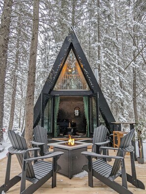 A cabin with a triangular roof, a central fire pit, and four Adirondack chairs. - The Hawthorn A-frame Cabin (Alberta Beach)