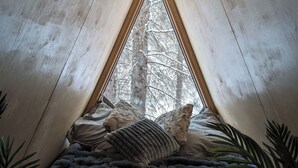 A cozy, triangular wooden tent with a bed at the base, surrounded by natural elements like trees and plants. - The Hawthorn A-frame Cabin (Alberta Beach)