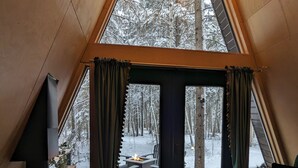 A cozy cabin interior with a wood-burning stove, a view of a snowy forest through large windows, and a wooden deck outside. - The Hawthorn A-frame Cabin (Alberta Beach)