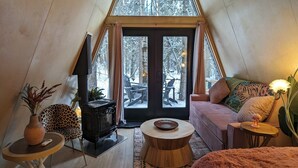 A cozy, modern loft bedroom with a sloped ceiling, a large window offering a view of the outdoors, a plush sofa, a round coffee table, and a fireplace. - The Fern A-frame Cabin (Alberta Beach)