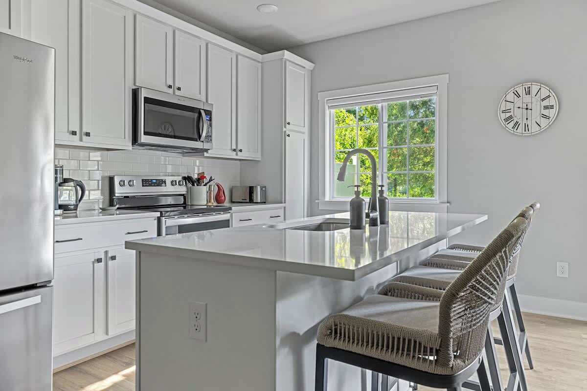 A modern kitchen with white cabinetry, a central island, bar stools, a microwave, an oven, and a large window with a view of greenery.