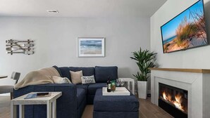Un salon moderne avec un canapé d’angle, une cheminée, un téléviseur et un tableau encadré représentant une plage.