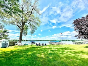 A grassy area with a large tree, a small shed, and a dock with boats.
