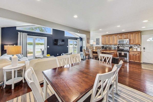 A spacious dining area with a large wooden table and white chairs, a kitchen with wooden cabinets and modern appliances, and a balcony with a view of the sea.