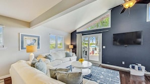 A modern living room with a sectional sofa, a round coffee table, a flat-screen TV, and a large window with blinds. - Shanahan Shores | Delavan Lakefront Cottage (Delavan)