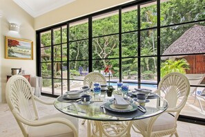 A dining area with a glass table, white wicker chairs, and a view of a swimming pool and lush greenery.