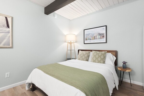 A bedroom with a bed, a bedside lamp, a framed picture on the wall, and a small table with a vase.