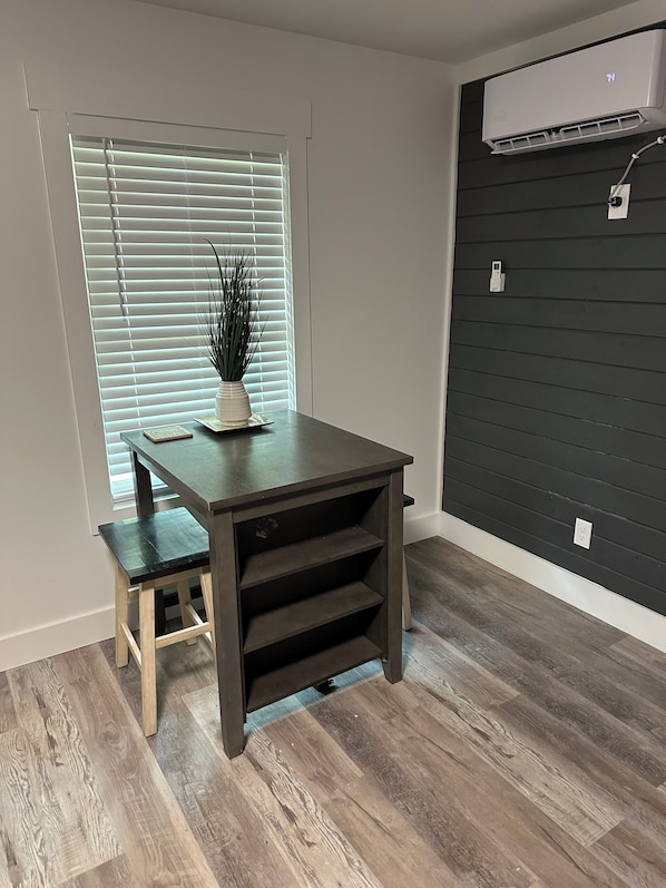 A room with a dark wooden table and bench, a potted plant, a window with blinds, and a wall-mounted air conditioner.